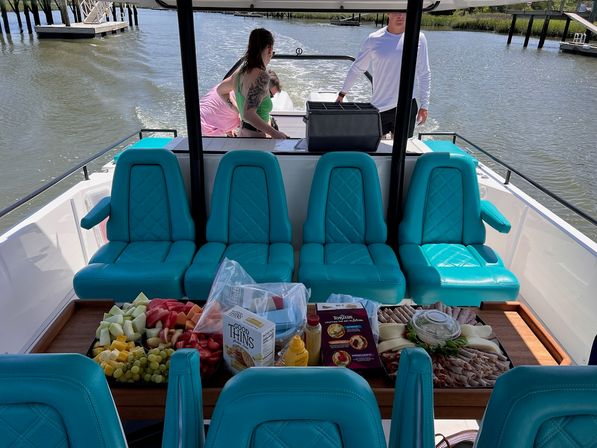 Boat cockpit with bright turquoise seats and a wooden table piled with fruit, cheese, charcuterie, crackers and condiments, people at the stern and a marina channel beyond.