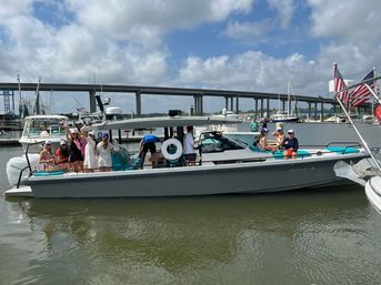 Group of people enjoying a sleek gray motorboat at a busy marina with sailboats and a concrete bridge in the background beneath a sunny, partly cloudy sky