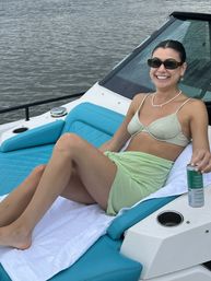 Smiling woman in a bikini top and light green sarong lounging on a turquoise cushioned boat seat, wearing sunglasses and a pearl necklace while holding a canned drink with calm water in the background.