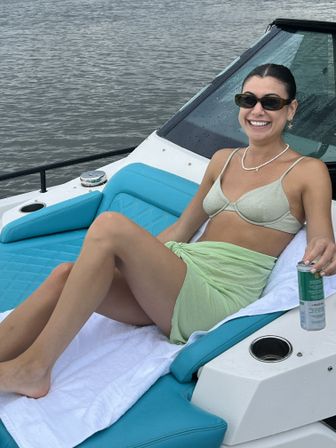 Smiling woman in a bikini top and light green sarong lounging on a turquoise cushioned boat seat, wearing sunglasses and a pearl necklace while holding a canned drink with calm water in the background.