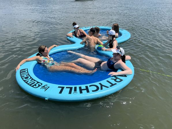 Friends lounging on linked blue inflatable floating pools in a calm lake, wearing swimsuits and caps and passing a small ball on a sunny summer day.