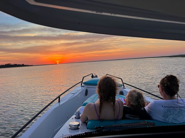 Three passengers on a motorboat bow watching a vivid orange sunset over calm coastal waters and marshy shoreline