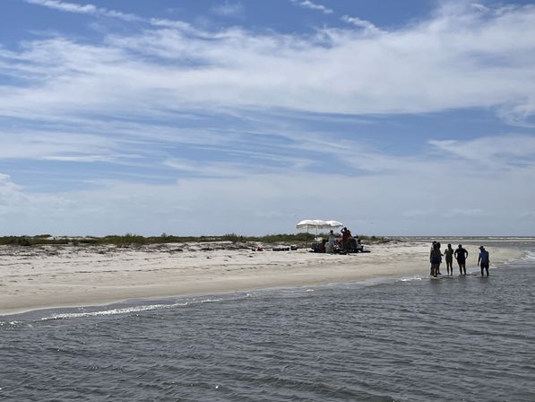 Sunny coastal sandbar with shallow water, a white beach umbrella and a small group of people walking along the shore under wispy blue clouds.