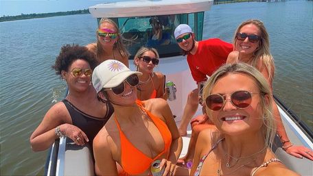 Seven friends smiling and posing on a sunlit motorboat in calm bay waters, wearing colorful swimsuits and sunglasses and holding canned drinks on a fun summer boating outing.