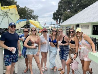 Smiling group of friends in summer outfits holding drinks on a sunny waterfront boardwalk, posing by a dockside bar with colorful umbrellas, wooden decking and water views — relaxed vacation vibe.