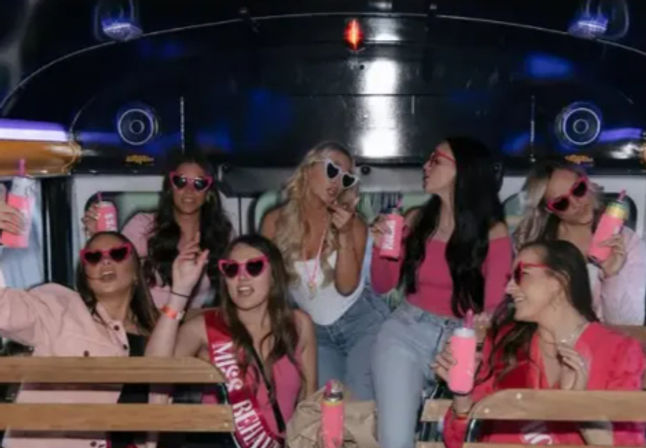 Group of women in pink sunglasses and sashes celebrating a bachelorette party on a party bus, holding pink drinks and posing under neon lights