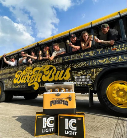 Friends leaning out of a black-and-yellow party bus, cheering with cans of light beer above a yellow cooler and stacked beer boxes on the pavement under a sunny blue sky.