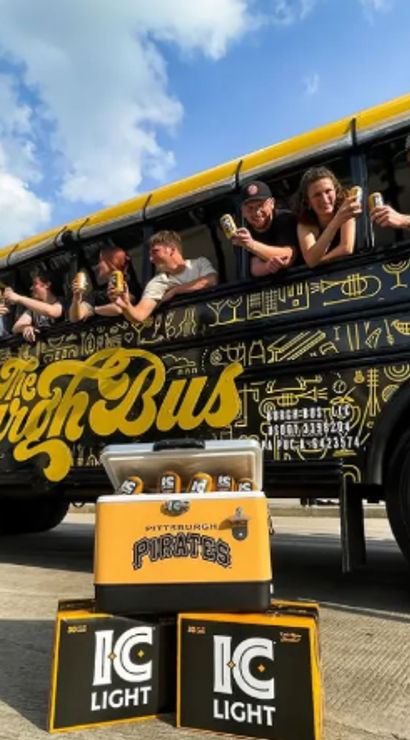 Friends leaning out of a black-and-yellow party bus, cheering with cans of light beer above a yellow cooler and stacked beer boxes on the pavement under a sunny blue sky.