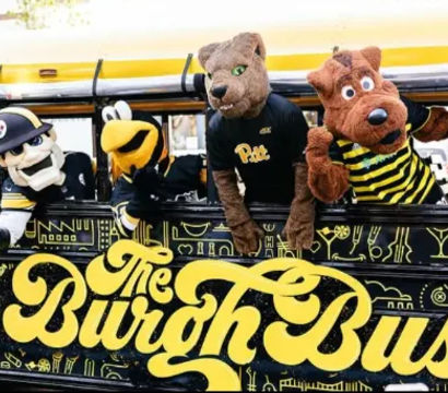 Yellow-and-black decorated bus with four playful sports mascots — a panther, a bee and two costumed characters — leaning from the windows in a festive street scene.