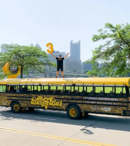 Person standing on roof of a yellow decorated tour bus holding a giant orange 3 balloon, Pittsburgh skyline and riverside park with green trees in the sunny background.