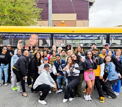 Large diverse group of people smiling and posing in front of a yellow party bus on a city street — friends on a playful urban group outing.