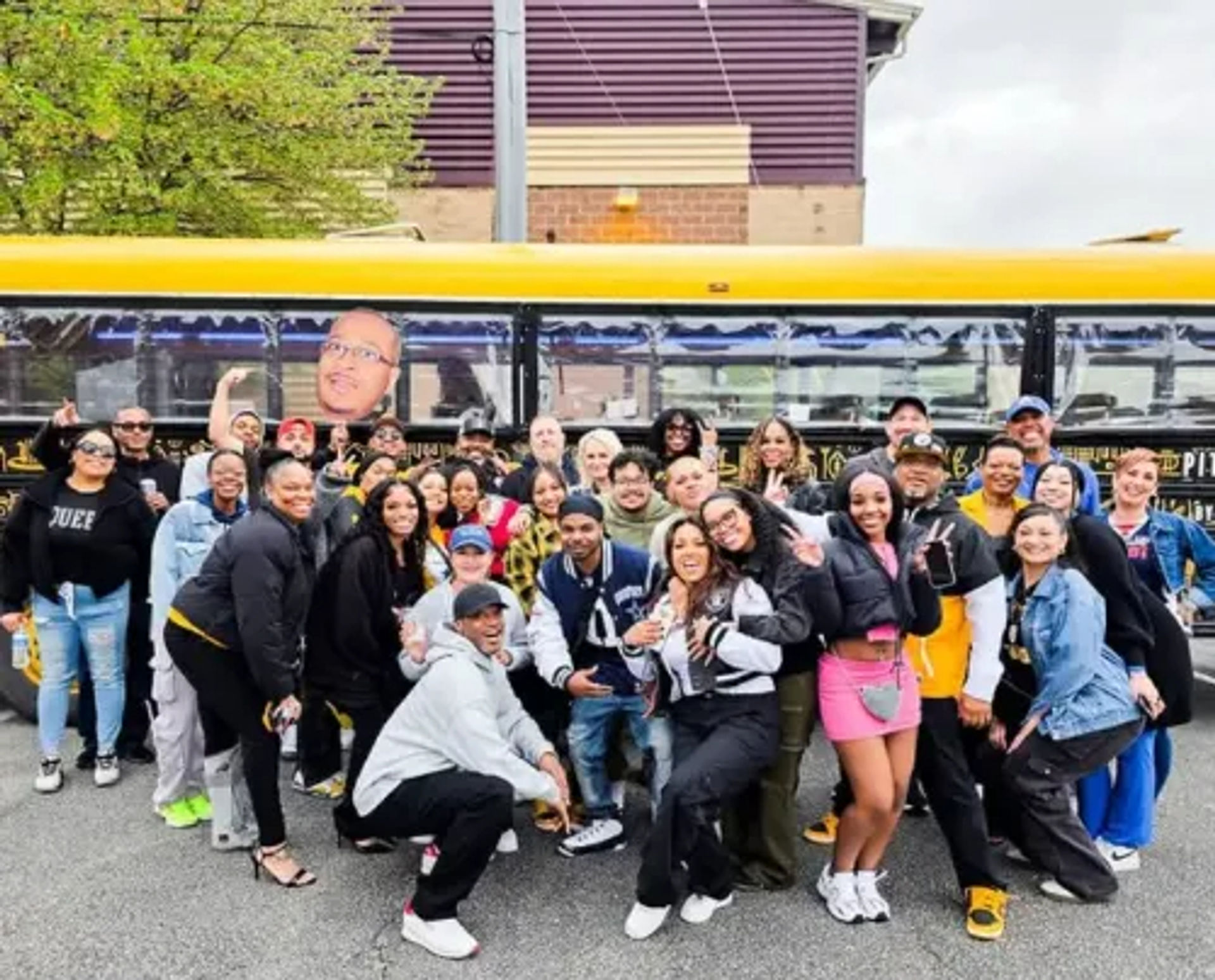 Large diverse group of people smiling and posing in front of a yellow party bus on a city street — friends on a playful urban group outing.