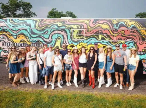 Group of young adults in summer outfits and cowboy boots smiling and posing in front of a colorful psychedelic tour bus with a swirly mural on a grassy outdoor lot