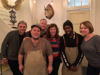 Group photo in a cozy restaurant dining room: seven smiling people including two apron-wearing chefs, decorative mounted animal head and warm pendant lights.
