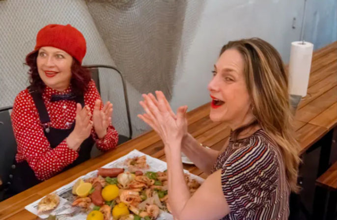 Two cheerful women clapping at a wooden counter in a casual eatery, sharing a seafood boil of shrimp, lemons and sausage served on butcher paper.