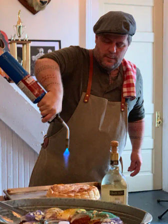 Chef in apron and flat cap using a blue-tipped culinary blowtorch to caramelize a puff pastry on a tray, bottle of sparkling wine nearby in a cozy farmhouse kitchen