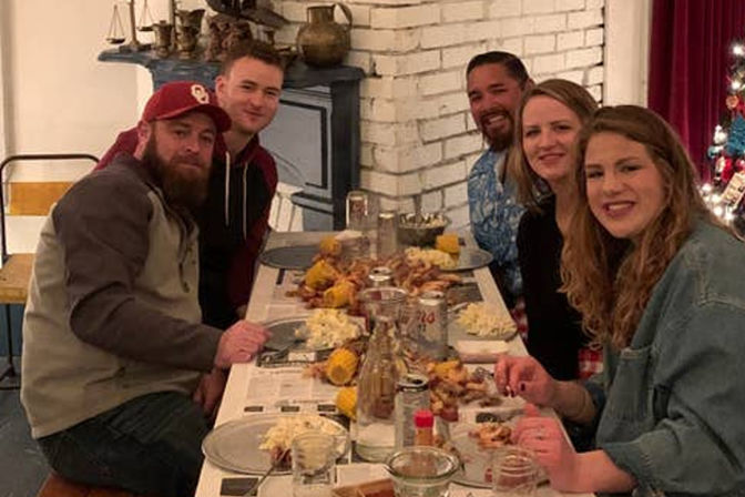 Five smiling friends at a long table in a cozy brick-walled room enjoying a casual seafood boil with corn, potatoes, and drinks.