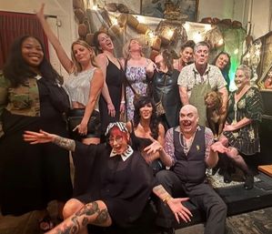 About a dozen adults and a dog in a playful group photo at a cozy indoor party, people laughing and striking fun poses in front of fishing-net and buoy décor with warm string lights.