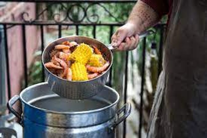 Hand holding a metal colander filled with cooked shrimp and corn on the cob over a large outdoor pot — backyard seafood boil.