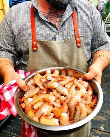 Person wearing an apron and patterned shirt holding a large metal bowl piled with fresh shrimp on a wooden deck, red-checkered towel and visible tattoos, outdoor seafood prep