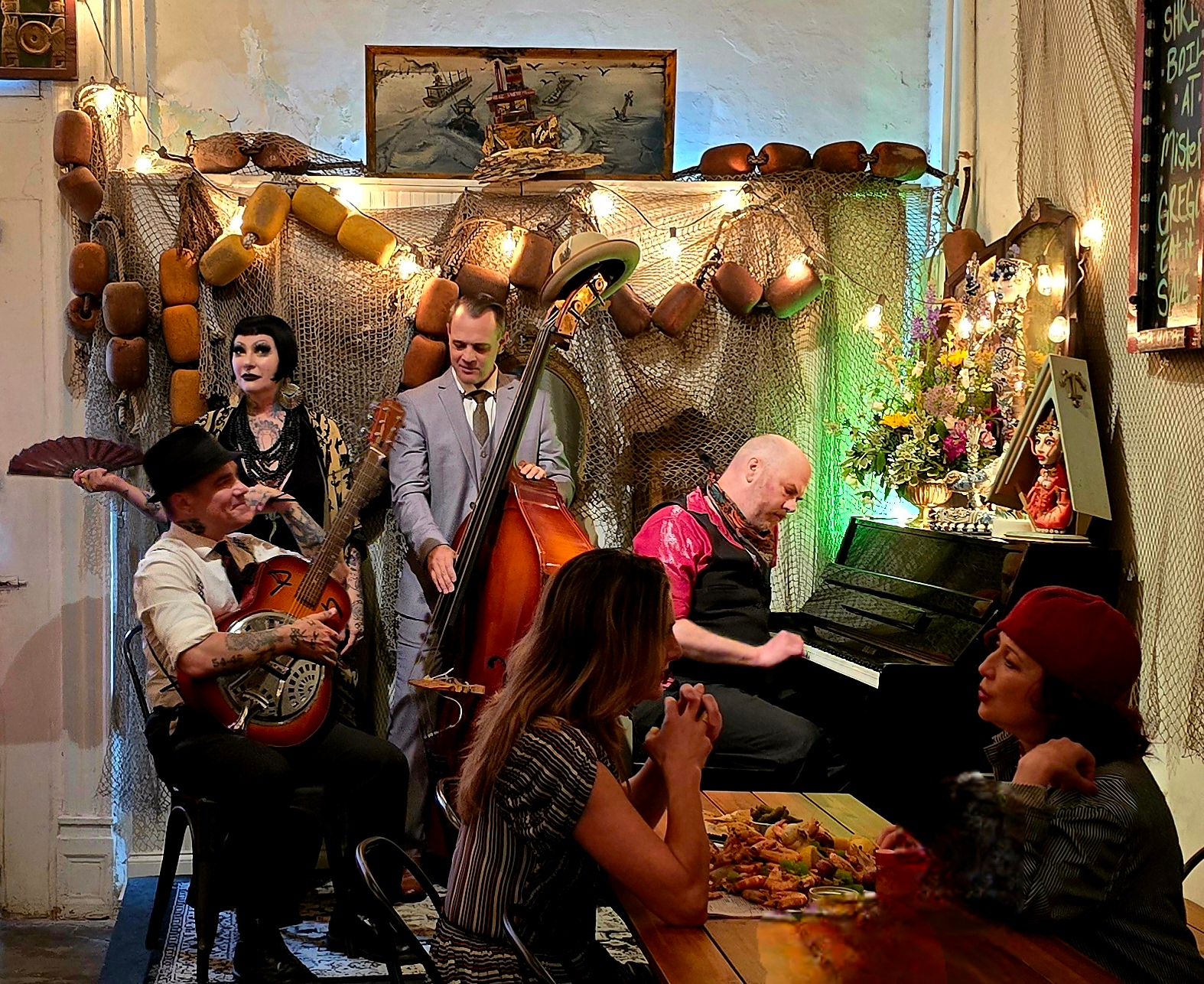 Intimate live music scene: jazz trio with upright bass, resonator guitar and pianist performing in a cozy, sea-themed cafe adorned with fishing nets, buoy decor, string lights and diners at a wooden table.