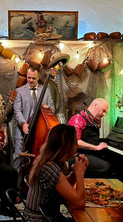 Intimate live music scene: jazz trio with upright bass, resonator guitar and pianist performing in a cozy, sea-themed cafe adorned with fishing nets, buoy decor, string lights and diners at a wooden table.