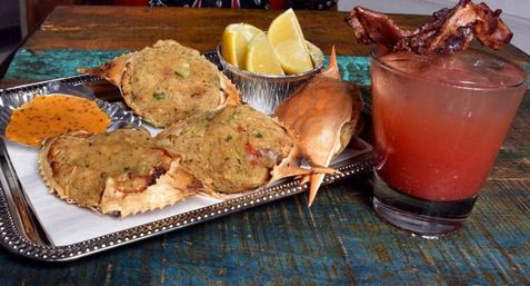 Baked stuffed crab shells on a silver tray with lemon wedges and dipping sauce, paired with a bacon-topped tomato cocktail on a rustic wooden table.