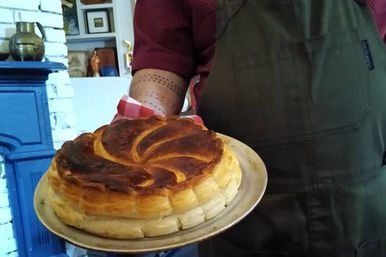 Person in an apron holding a golden, flaky spiral puff pastry tart on a round metal baking tray in a cozy kitchen with a blue mantel