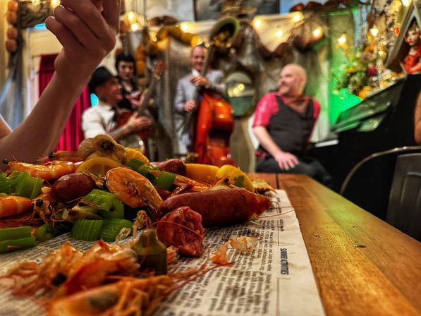 New Orleans-style seafood boil — shrimp, crawfish, sausage, celery and lemon on a newspaper-lined wooden table, with a blurred live jazz trio (upright bass and piano) in a cozy, festive bar