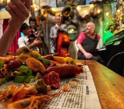 New Orleans-style seafood boil — shrimp, crawfish, sausage, celery and lemon on a newspaper-lined wooden table, with a blurred live jazz trio (upright bass and piano) in a cozy, festive bar