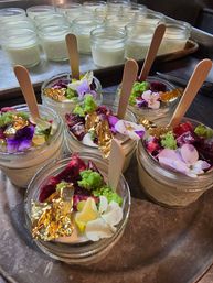 Gourmet mini dessert jars in glass with wooden spoons, topped with edible flowers, green sponge crumbs, berry compote and gold leaf, arranged on a metal tray.