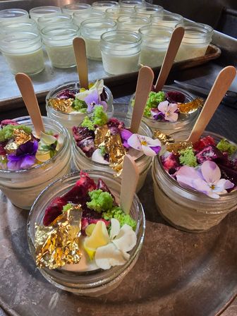 Gourmet mini dessert jars in glass with wooden spoons, topped with edible flowers, green sponge crumbs, berry compote and gold leaf, arranged on a metal tray.