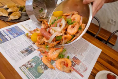 Hands pouring a coastal-style shrimp boil from a metal bowl — whole shrimp, smoked sausage, lemon wedges, sliced onions and green peppers spilling onto newspaper-lined wooden table, with breadcrumb crab shells and dipping sauce at the edge.