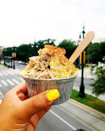 Hand with bright yellow nail polish holding an aluminum to-go cup of creamy mac and cheese topped with shredded crab and crispy breadcrumbs, wooden spoon inserted, blurred city street and trees in the background.