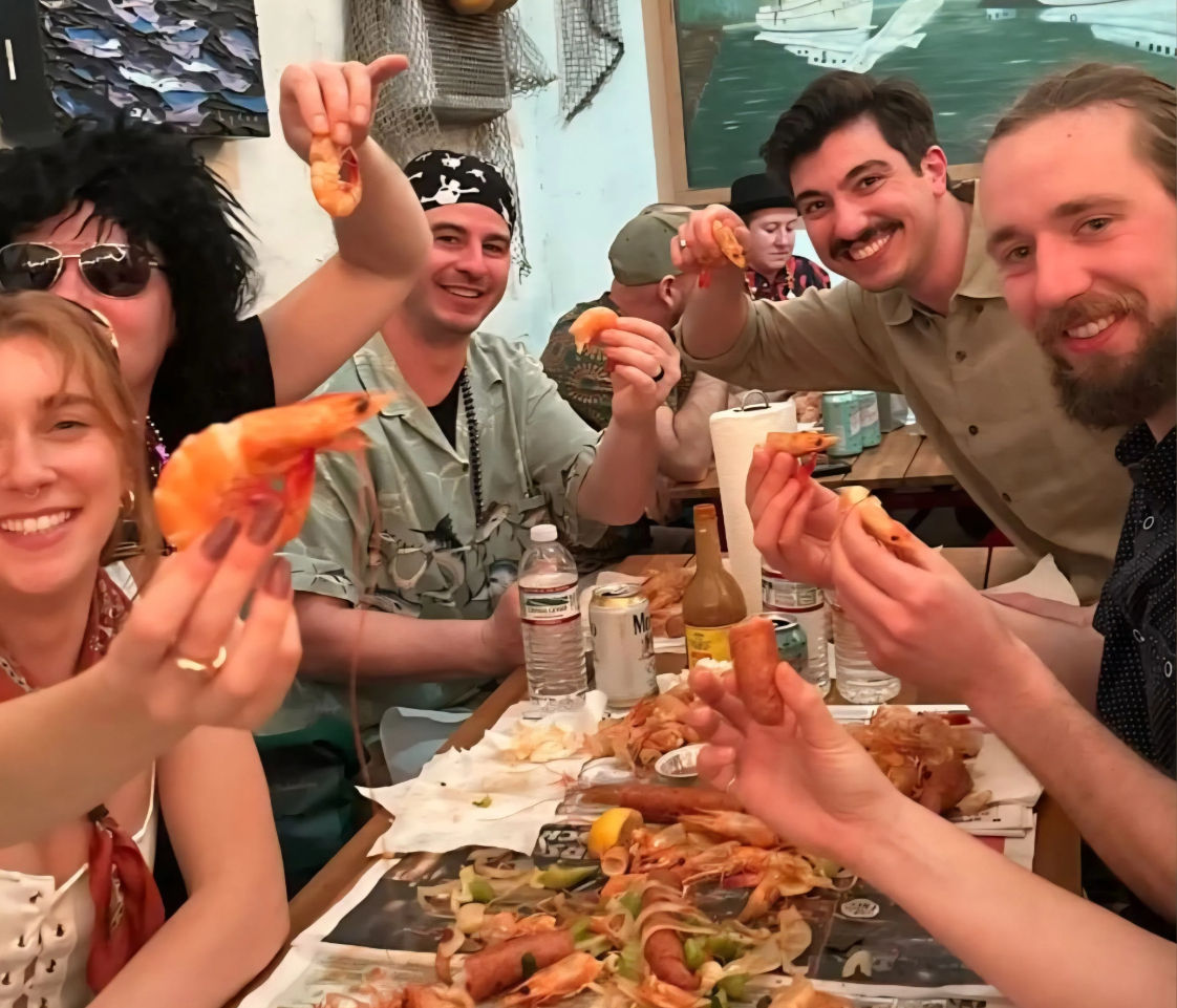 Smiling group at a casual seafood restaurant enjoying a shrimp boil — holding peel-and-eat shrimp over a paper-lined communal table with shells, lemon wedges and drinks.