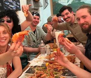 Smiling group at a casual seafood restaurant enjoying a shrimp boil — holding peel-and-eat shrimp over a paper-lined communal table with shells, lemon wedges and drinks.