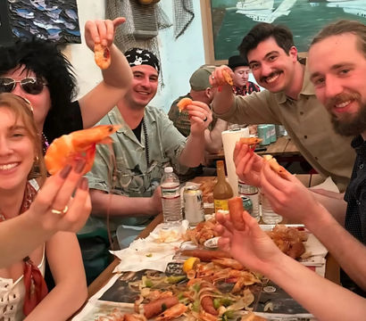 Smiling group at a casual seafood restaurant enjoying a shrimp boil — holding peel-and-eat shrimp over a paper-lined communal table with shells, lemon wedges and drinks.