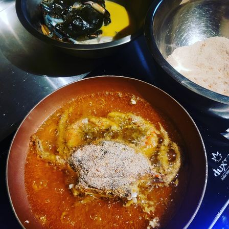 Sizzling breaded soft-shell crab frying in hot oil in a skillet, with metal bowls of egg wash and breadcrumbs on a stainless-steel kitchen surface.