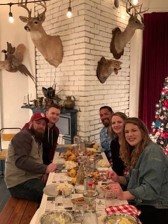 Five people enjoying a casual holiday seafood boil at a long rustic table in a cozy indoor dining room with a white painted brick corner wall displaying mounted deer, fox and pheasant heads, string lights, and a decorated Christmas tree.