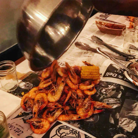 Casual seafood boil scene with a pile of seasoned boiled shrimp and corn on the cob being tipped from a metal pot onto a newspaper-covered table with tongs and mason jars nearby