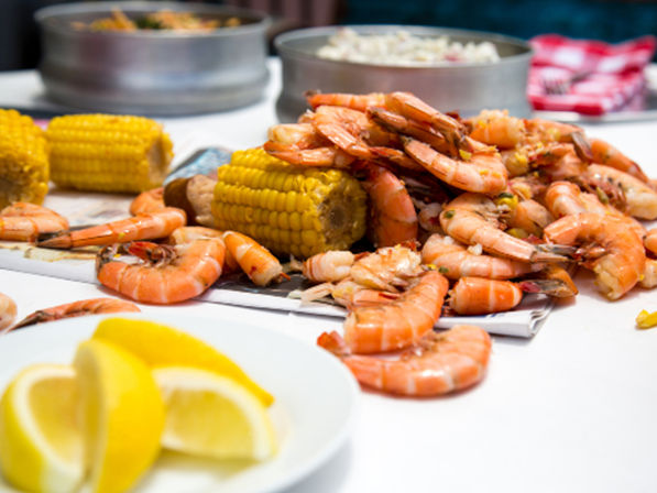 Close-up of a seafood boil: piled cooked shrimp and corn on the cob on a table with lemon wedges and metal serving bowls
