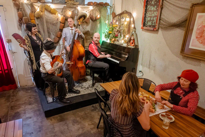 Toe-tapping jazz quartet (upright bass, piano, resonator guitar, singer) performing on a tiny stage with vintage nautical decor while two diners share a meal at a cozy neighborhood cafe.