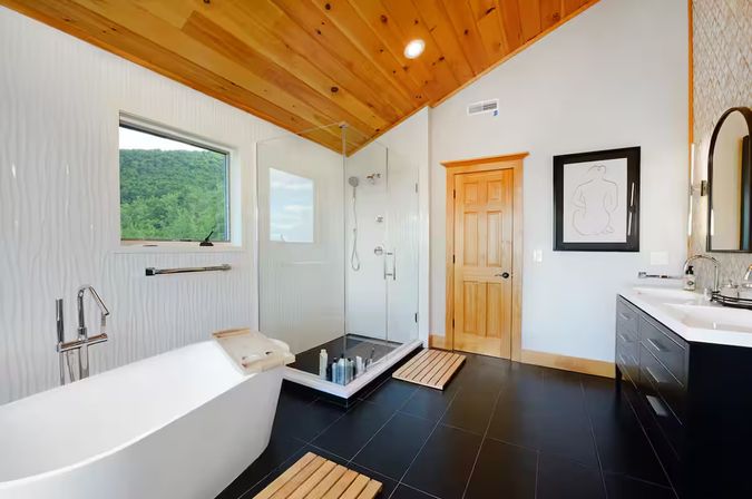 Modern mountain-view bathroom with a freestanding white tub, glass walk-in shower, black tile floor, wood-paneled vaulted ceiling and double vanity.