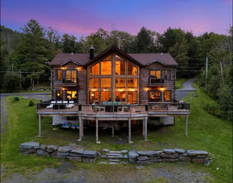 Cozy rustic log cabin lodge at dusk with glowing floor-to-ceiling windows, wide wooden deck with outdoor seating, kayaks stored under the deck, stone retaining wall and grassy lawn surrounded by forest beneath a purple sunset sky.