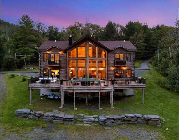 Cozy rustic log cabin lodge at dusk with glowing floor-to-ceiling windows, wide wooden deck with outdoor seating, kayaks stored under the deck, stone retaining wall and grassy lawn surrounded by forest beneath a purple sunset sky.