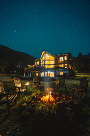 Inviting illuminated mountain cabin at night under a star-filled sky, wooden deck and glowing fire pit surrounded by Adirondack chairs on a grassy lawn.