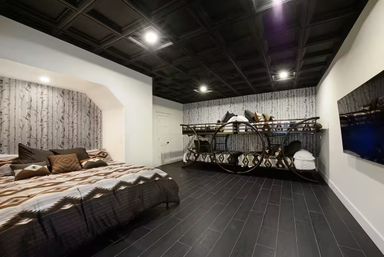 Basement bedroom with large bed and circular-frame bunk beds, black coffered ceiling, dark wood-look tile floor, birch-pattern wallpaper accent wall and wall-mounted TV.