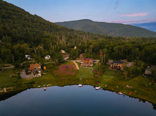 Aerial view of lakefront cabins and docks with colorful kayaks on a calm mountain lake at dusk, surrounded by green forested hills and a wildflower meadow.