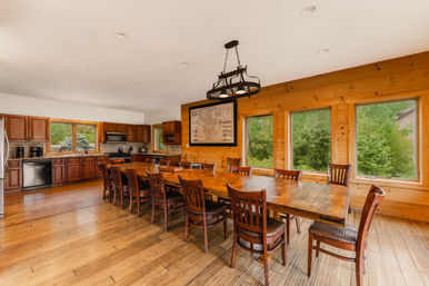 Open-concept wood-paneled dining room and kitchen with a long farmhouse table seating twelve, rustic chandelier, hardwood floors, and large windows overlooking lush green trees.