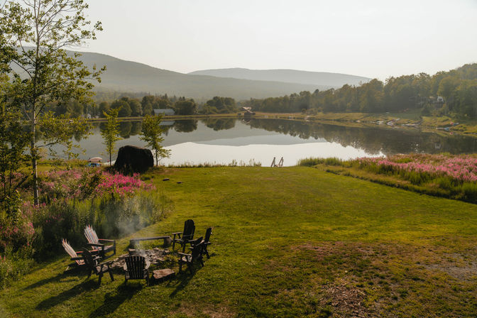 Cozy lakefront scene with Adirondack chairs around a smoldering fire pit on a grassy meadow edged by pink wildflowers, a calm reflective lake with a small dock and tree-covered hills in soft morning light.